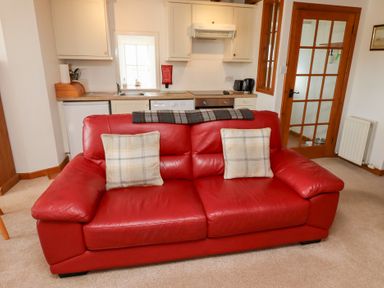 A red leather sofa with two plaid cushions in a living space with a small kitchen area and a wooden door at The Bothy Lowick near Holy Island