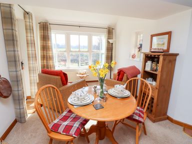 A dining area with a round wooden table set for two, wooden chairs with plaid cushions, wicker armchairs with red cushions, a wooden cabinet with tea jars, and large windows with plaid curtains at The Bothy in Lowick near Holy Island
