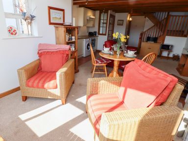 A living room with wicker armchairs, a wooden dining table set, kitchen area, and staircase at The Bothy in Lowick near Holy Island