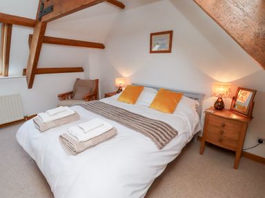 A bedroom with a double bed with white linens yellow pillows and folded towels a wooden chair bedside tables with lamps and wood ceiling beams at The Bothy Lowick near Holy Island