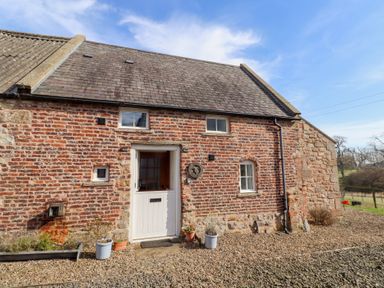 A brick cottage with a half door and three windows and potted plants on a gravel surface at The Bothy in Lowick near Holy Island