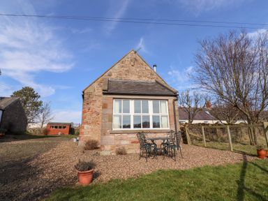 A stone house with a large window a black metal table and chairs on gravel outside at The Bothy in Lowick near Holy Island