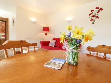 A dining table with flowers and a guide book with a red sofa and lamp in the background at Poppy Cottage Little Glemham near Wickham Market