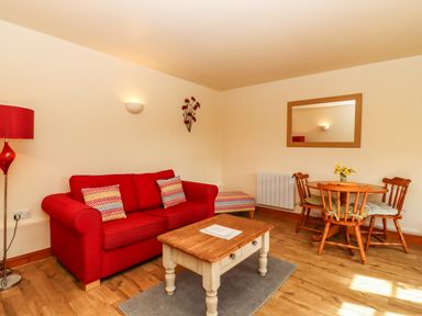 A living room with a red sofa two patterned cushions a wooden coffee table and a round dining table with four chairs at Poppy Cottage in Little Glemham near Wickham Market
