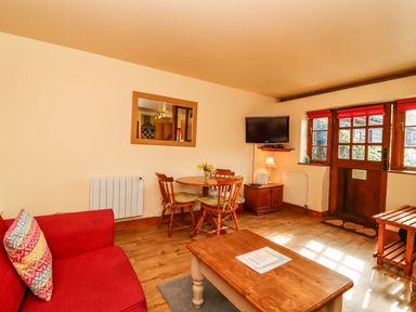 A living room with a red sofa a wooden coffee table a round dining table with chairs a wall-mounted television and wooden door with windows at Poppy Cottage in Little Glemham near Wickham Market