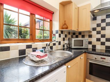 A kitchen with a stainless steel sink and drying rack black and white tiled walls wooden cabinets microwave and oven at Poppy Cottage in Little Glemham near Wickham Market