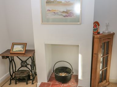 A small fireplace with a black pot filled with stones inside a living room with a wooden cabinet and a sewing machine table at 28 Oxborough in Oxborough