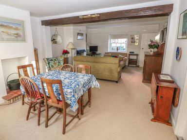A dining area with a wooden table covered by a blue and white tablecloth and four chairs next to a living area with sofas a TV and wooden furniture at 28 Oxborough in Oxborough