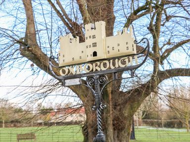 A village sign with a castle and the word Oxborough in a park with trees and a bench at 28 Oxborough in Oxborough