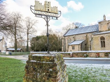 A village sign on a stone base with a church and trees in the background at 28 Oxborough in Oxborough