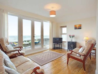 A living room with wooden armchairs a rug a television and large glass doors overlooking the sea at Trysor Y Mor in Borth
