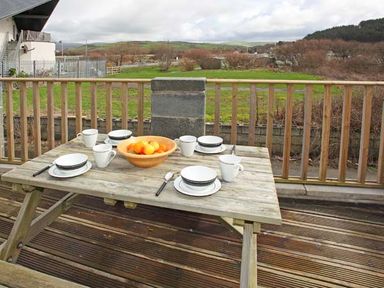 An outdoor wooden picnic table set with mugs plates and a bowl of fruit on a deck with a wooden railing and a grassy field in the background at Trysor Y Mor in Borth