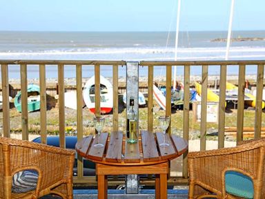 A wooden balcony with two wicker chairs and a wooden table with a bottle and two glasses overlooking boats on a beach at Trysor Y Mor in Borth