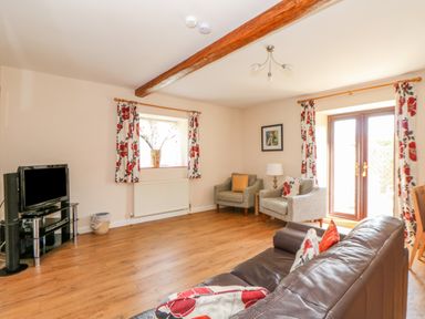 A living room with a brown sofa, two armchairs, a TV on a stand, floral curtains, and wooden floor at The Granary Cottage in Gayton near King's Lynn