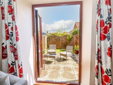 A patio with chairs and a table outside a door with floral curtains at The Granary Cottage in Gayton near King's Lynn