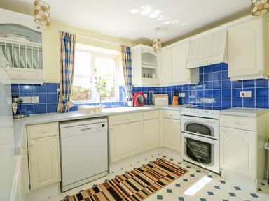 A kitchen with white cabinets blue tile backsplash a dishwasher stove and window with check curtains at The Granary Cottage in Gayton near Kings Lynn