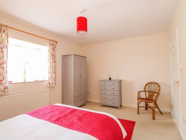 A bedroom with a bed covered by a white and red blanket two grey dressers a wicker chair and a window with floral curtains at The Granary Cottage in Gayton near King's Lynn