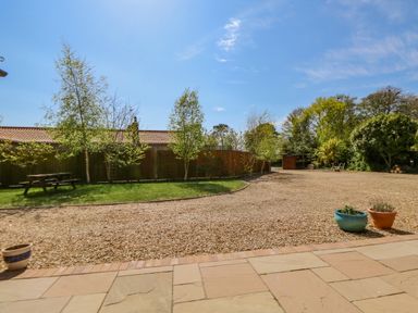 An outdoor area with a gravel driveway, trees along a wooden fence, grass patch with a picnic table, and potted plants on a stone patio at The Granary Cottage in Gayton near Kings Lynn