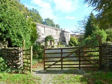 A stone cottage behind a wooden gate and stone walls surrounded by trees and greenery at High Kiln Bank Cottage in Seathwaite Duddon Valley