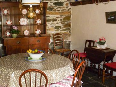 A dining room with a round table covered with a polka dot tablecloth surrounded by wooden chairs a wooden cabinet with plates and a potted plant and a small wooden table with a flower arrangement at High Kiln Bank Cottage Seathwaite Duddon Valley