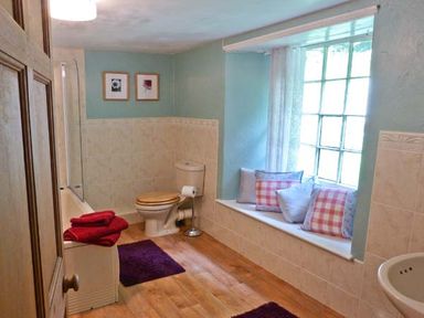 A bathroom with a toilet wooden seat a bath with red towels two framed pictures on the wall and a window seat with cushions at High Kiln Bank Cottage in Seathwaite Duddon Valley