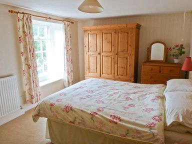 A bedroom with a bed covered in floral bedding next to a window with floral curtains and wooden furniture at High Kiln Bank Cottage in Seathwaite Duddon Valley