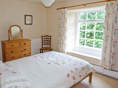 A bedroom with a bed covered in floral bedding a wooden dresser with a mirror a wooden chair and a window with floral curtains at High Kiln Bank Cottage in Seathwaite Duddon Valley
