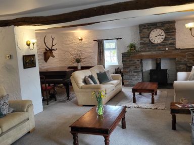 A living room with beige sofas wooden coffee tables a stone fireplace with a clock a mounted deer head and a piano at High Kiln Bank Cottage in Seathwaite Duddon Valley