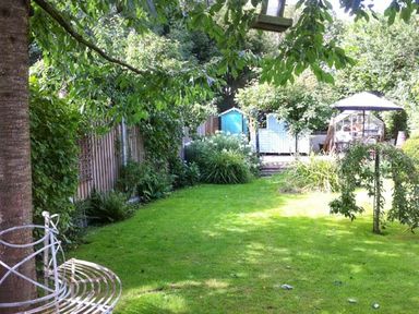 A garden with green grass green shrubs and trees a wooden fence a bird feeder a garden bench a shed and a patio umbrella at Teds Place in Aylsham
