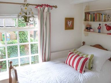 A bedroom with a single bed two striped pillows a window with curtains a chandelier a bookshelf with books and toys and a framed picture on the wall at Teds Place in Aylsham