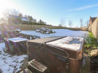 An outdoor area with a hot tub partially covered in snow a table with six chairs and a fenced grassy yard with some snow at Blencathra Barn in Penrith