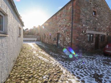 A cobblestone driveway between a white building and a stone building with snow on the ground and a red car parked at Blencathra Barn in Penrith