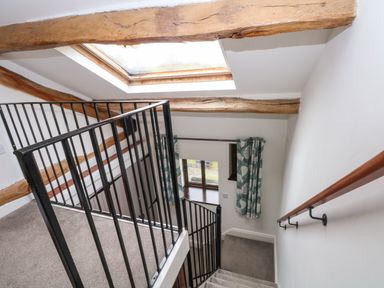 A staircase with black metal railing and wooden handrail under a skylight window with exposed wooden beams and a window with patterned curtains at Blencathra Barn in Penrith