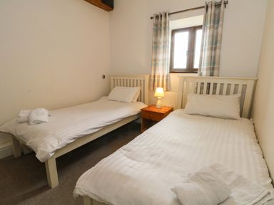 A bedroom with two single beds white bedding a wooden nightstand with a lamp and a window with curtains at Blencathra Barn in Penrith