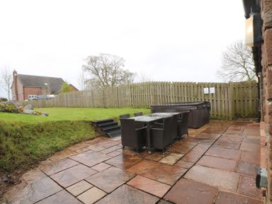 An outdoor patio area with a glass table and six wicker chairs next to a hot tub and grass lawn enclosed by a wooden fence at Blencathra Barn in Penrith