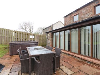 An outdoor patio with a glass table and six chairs next to a fenced yard and a hot tub at Blencathra Barn in Penrith