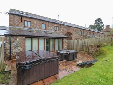 An outdoor patio area with a hot tub and dining table with chairs next to a stone house at Blencathra Barn in Penrith