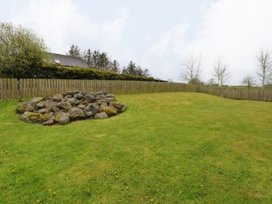 A grassy yard with a pile of rocks and a wooden fence with trees and a house in the background at Blencathra Barn in Penrith