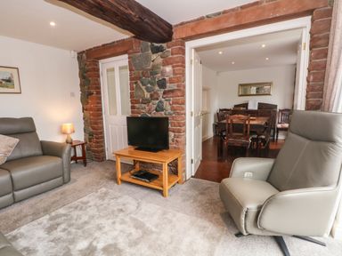 A living room with a grey armchair a sofa and a wooden TV stand with a television next to a dining room with wooden chairs at Saddleback Barn Newton Reigny near Penrith