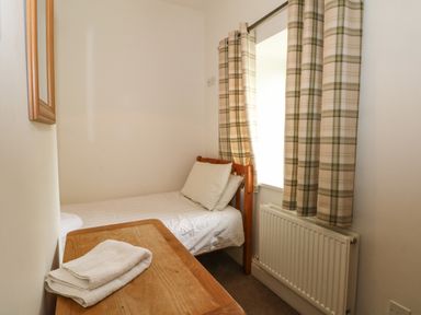A small bedroom with a single wooden bed with white linens beside a window with plaid curtains and a wooden desk with folded towels at Saddleback Barn in Newton Reigny near Penrith