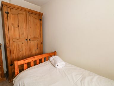 A bedroom with a wooden bed frame, a white bedcover, a rolled white towel on the bed, and a wooden wardrobe against the wall at Saddleback Barn in Newton Reigny near Penrith