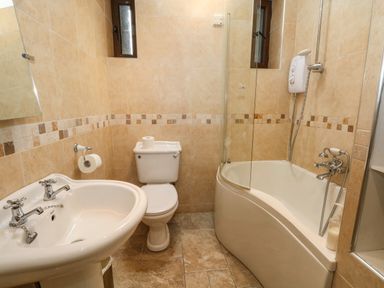 A bathroom with a white sink, toilet, curved bathtub with shower and beige tiled walls at Saddleback Barn in Newton Reigny near Penrith