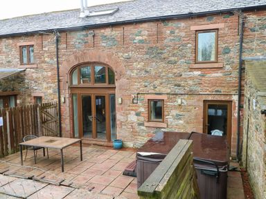 A patio with a table and chair hot tub and stone building with doors and windows at Saddleback Barn in Newton Reigny near Penrith