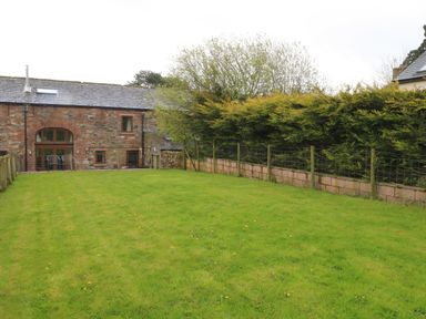 A fenced grass lawn with a stone building and trees in the background at Saddleback Barn in Newton Reigny near Penrith