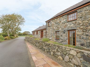 Stone cottage with wooden framed windows and door beside a paved walkway and road with trees in the background at Lily Cottage in Caeathro near Caernarfon