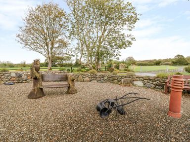 An outdoor area with a wooden bench carved with an owl next to a stone wall and trees at Lavender Cottage in Caeathro near Caernarfon