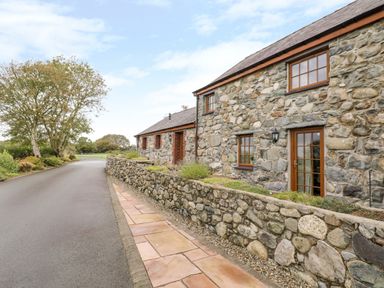 A stone house with wooden framed windows and a stone wall along a paved road at Lavender Cottage in Caeathro near Caernarfon