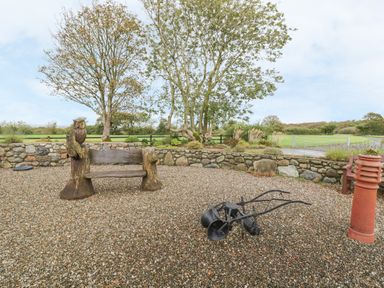 An outdoor area with a stone wall trees a wooden bench with owl carvings and old farming equipment at Bluebell Cottage in Caeathro near Caernarfon