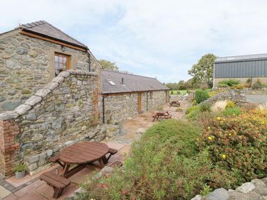A stone building with wooden picnic tables on a paved patio and garden bushes at Bluebell Cottage in Caeathro near Caernarfon
