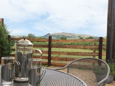 A metal outdoor table with glass and metal coffee pot and cups next to a wooden fence with hills in the background at Bicton Lodge in Clun